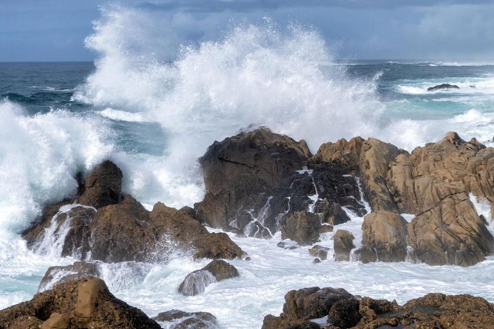 At Weston Beach in Point Lobos State Natural Reserve, powerful waves crash against jagged rock formations along the shoreline, sending white spray high into the air as turquoise water churns around the rugged coast.